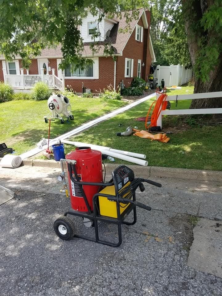 A red and black portable machine sits on a gravel driveway in front of a brick house, surrounded by construction materials and tools.