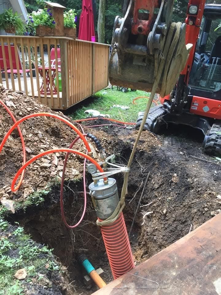 A close-up view of a construction site showing an excavated hole with plumbing equipment and orange hoses, alongside a backhoe..