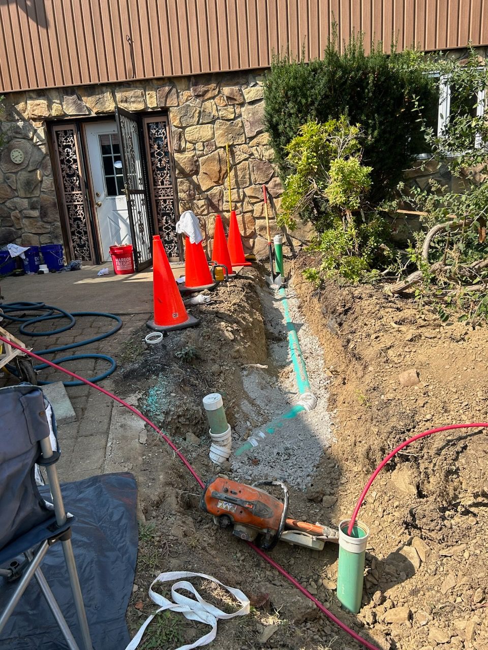 A trench with exposed pipes and tools outside a house, surrounded by orange cones and construction materials.