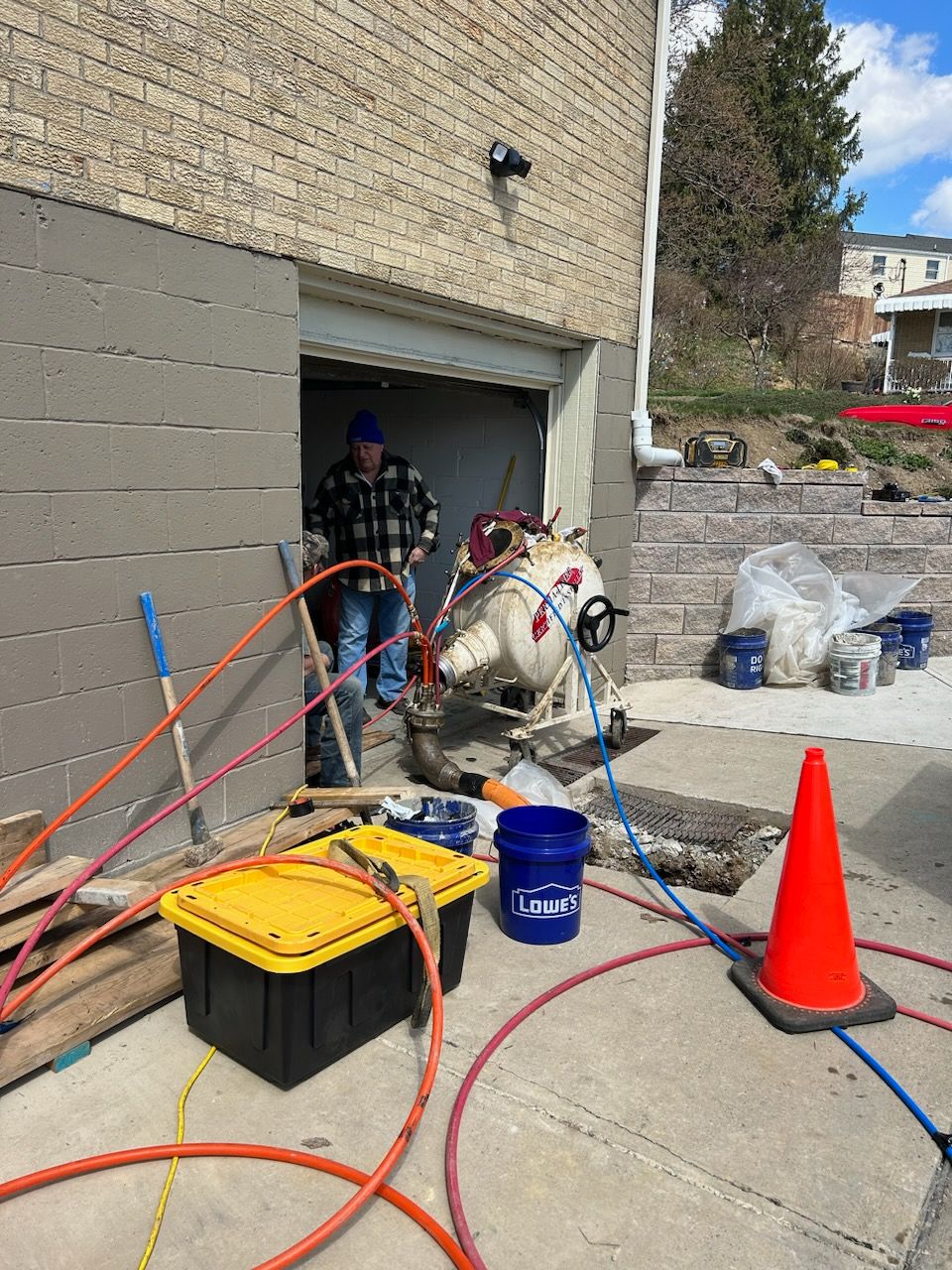 A worker stands at a garage entrance surrounded by construction equipment, hoses, and tools on a sunny day. Safety cone nearby.