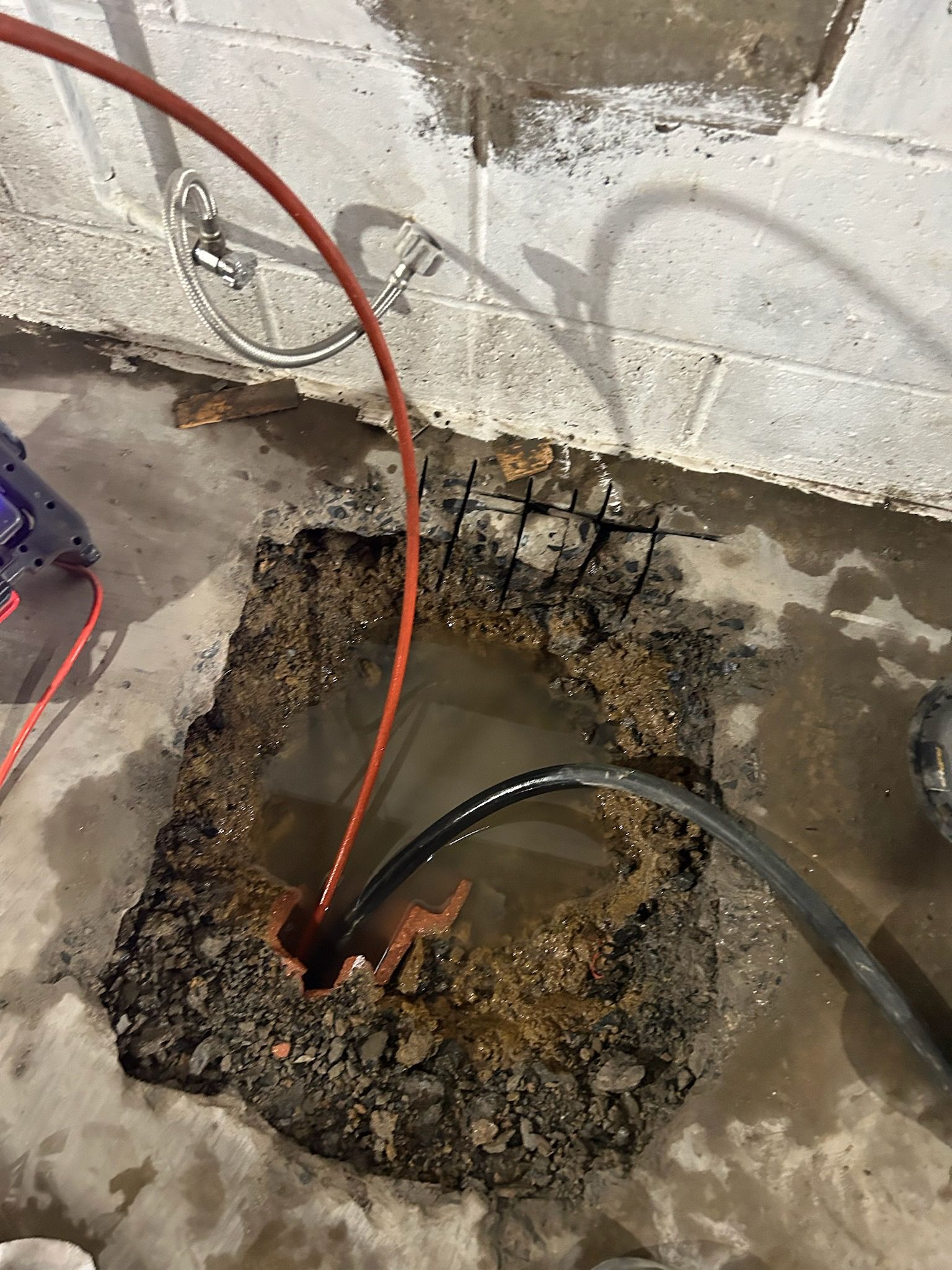 A deep hole filled with water in a basement, featuring red and black hoses linked to plumbing equipment against a concrete wall.