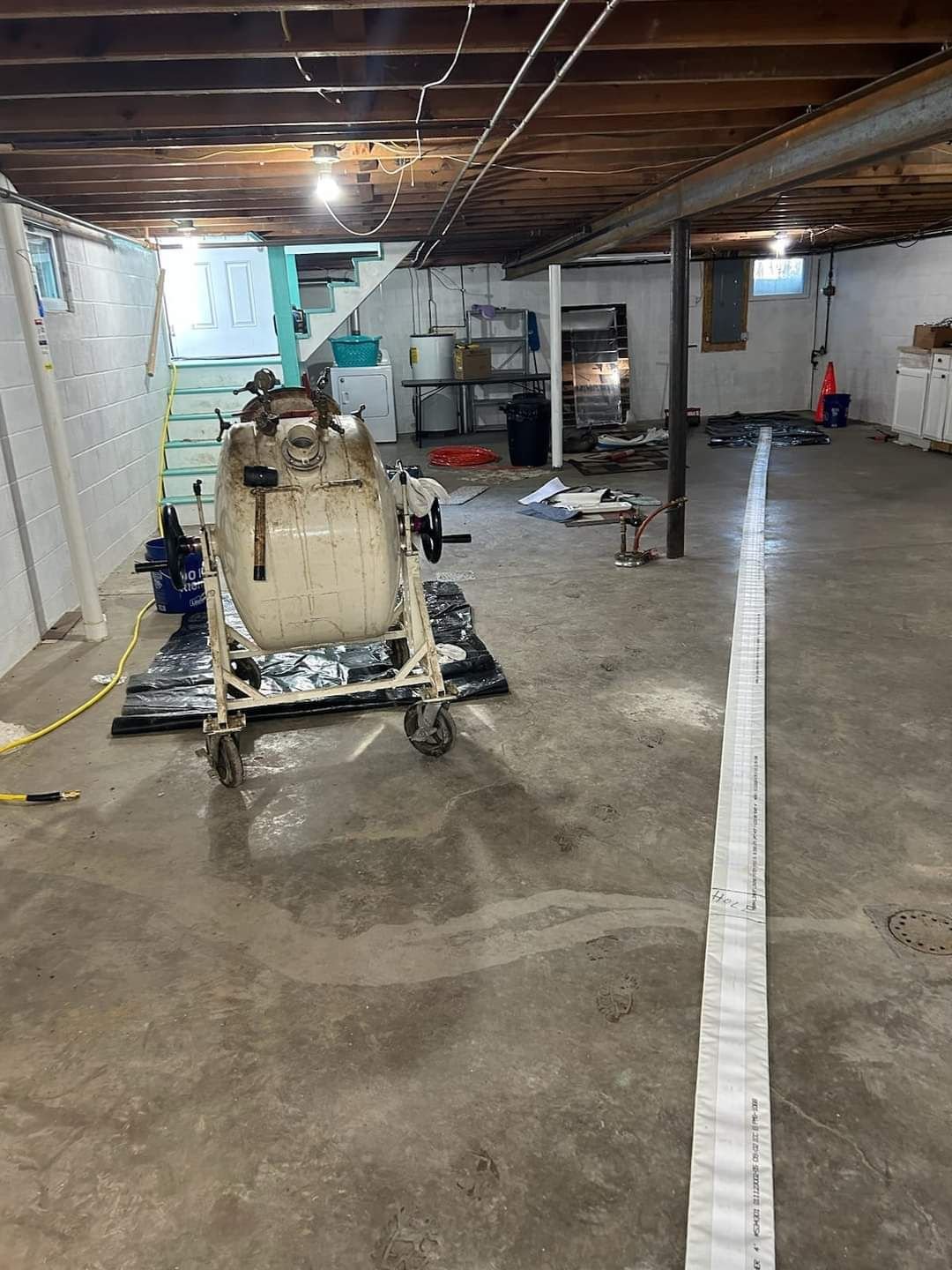 A spacious basement with a vintage air compressor on wheels, stairs leading up, and construction materials scattered across the concrete floor.