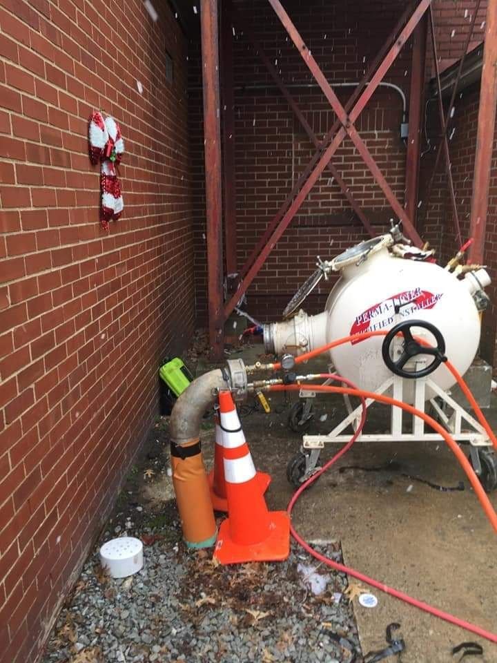 A snow-covered outdoor scene featuring a white pressure tank, colorful traffic cones, and a festive candy cane decoration on a brick wall.