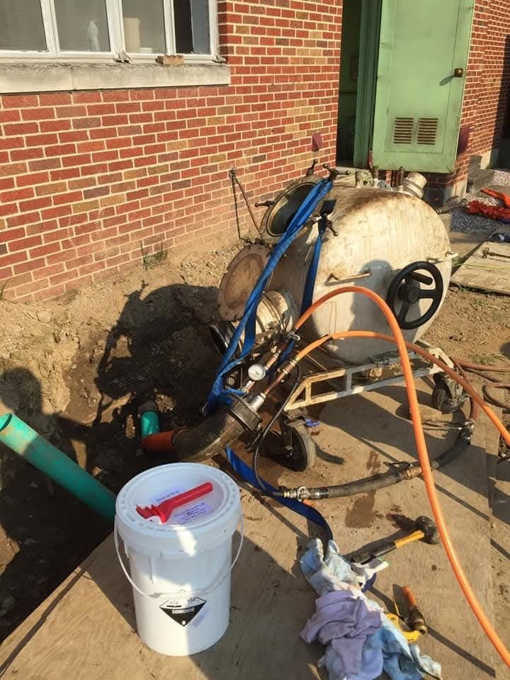 A construction site showing a variety of pipes and hoses, a white bucket with a red handle, and tools on a wooden surface.