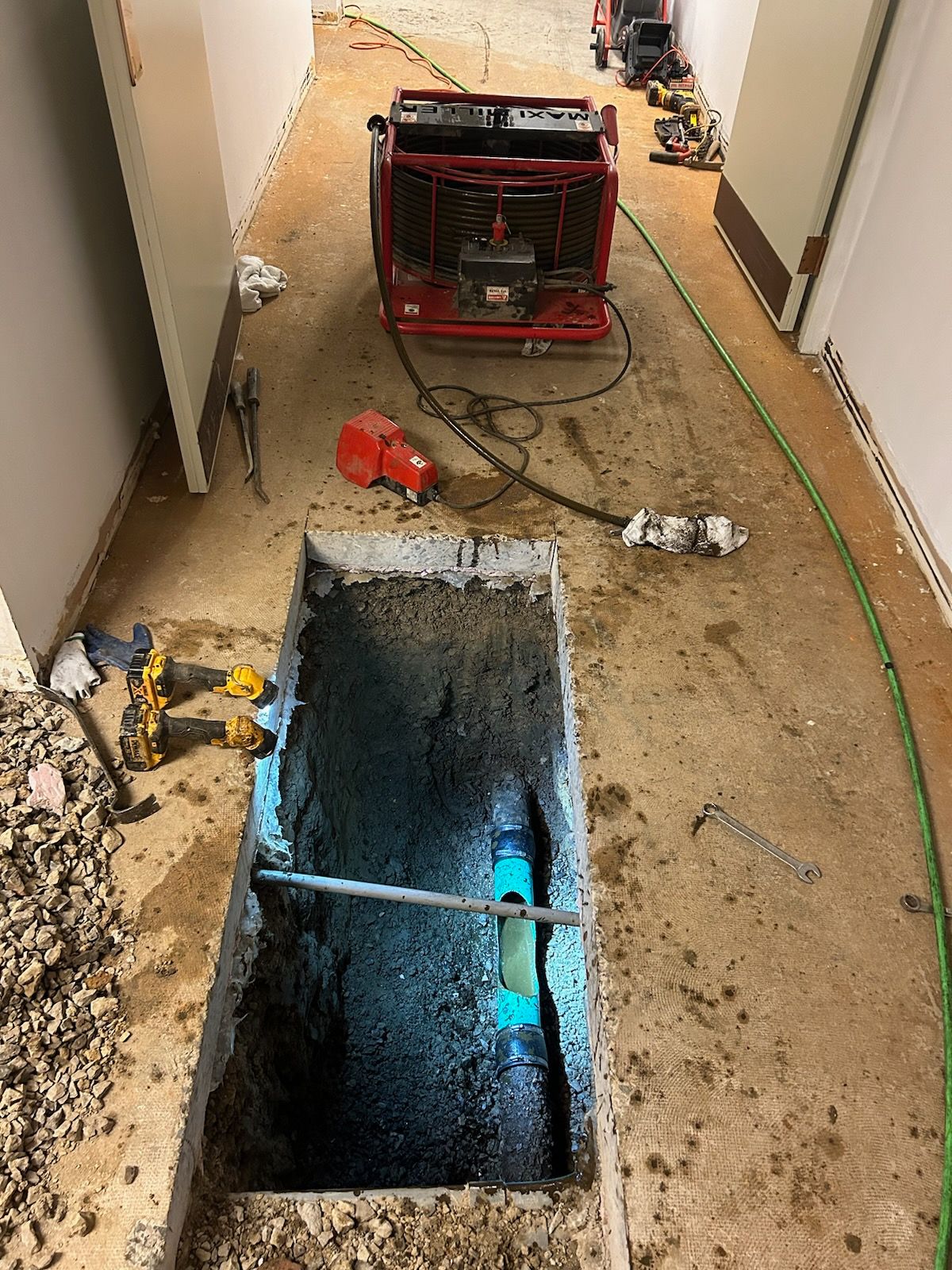 A concrete trench reveals a blue water pipe, surrounded by tools and a fan, in a construction corridor.