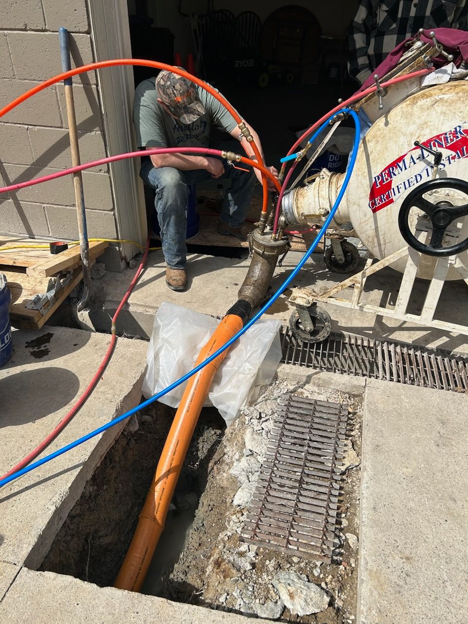 A worker connects colorful hoses to a machine beside an open excavation, with an orange pipe extending into the ground.